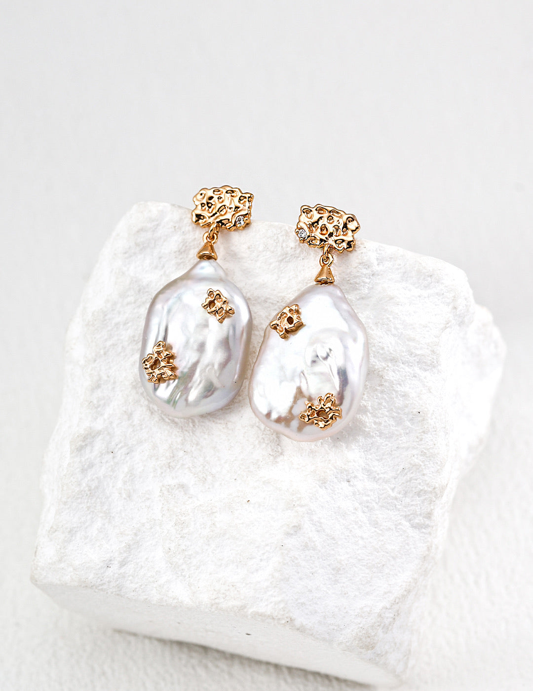 Mira Aura baroque pearl pendant earrings with gold floral accents on textured white stone