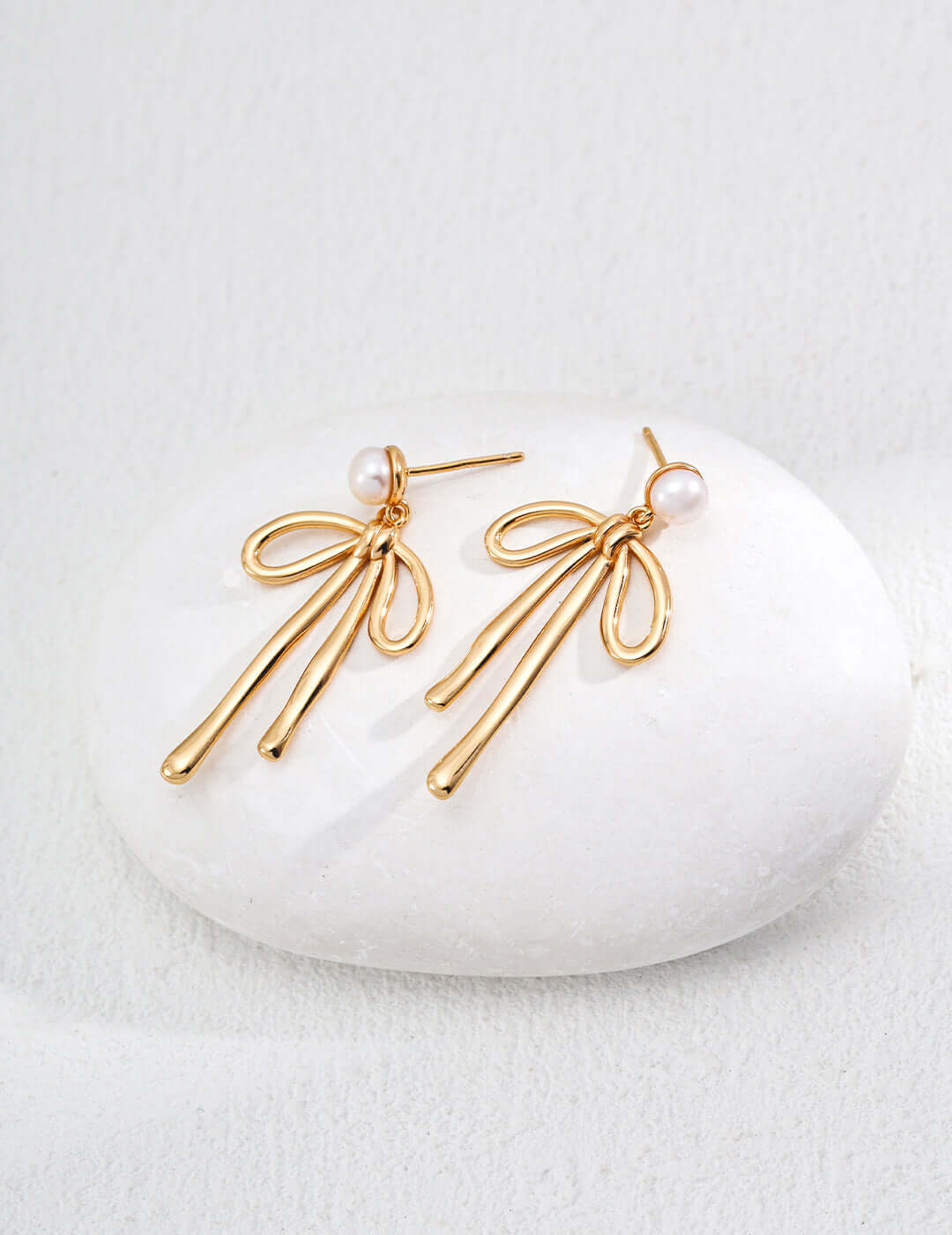 Mira Aura modern gold dangle earrings with pearl studs and ribbon like loops on white stone display