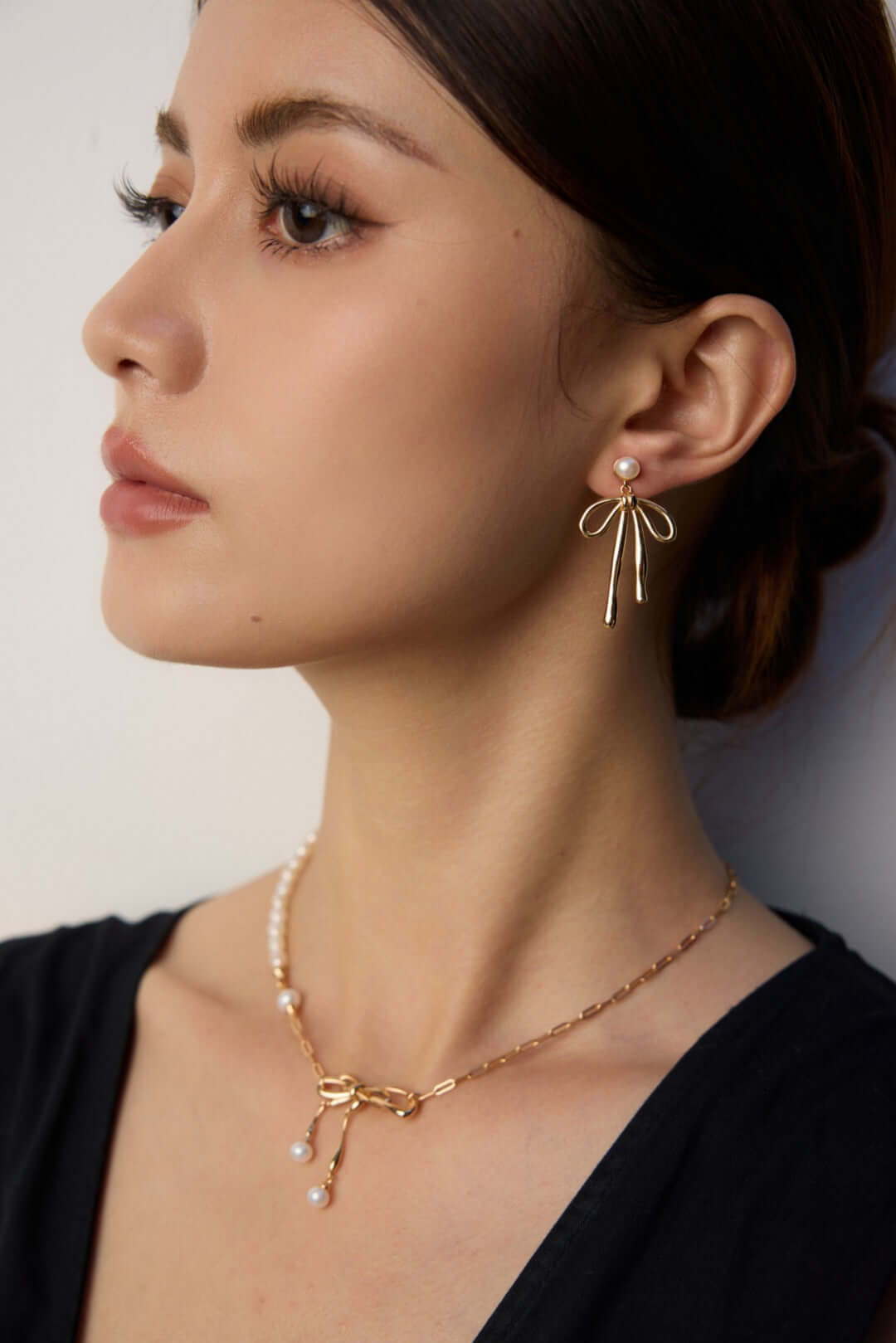 Woman wearing modern gold dangle earrings with pearl stud and delicate bow ribbon design