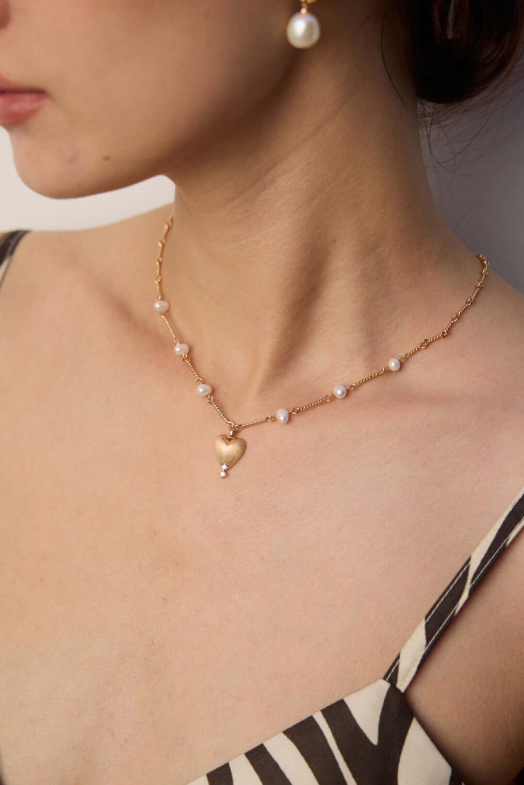 Model wearing gold plated heart pendant necklace with pearl station chain resting on collarbone and matching pearl earring